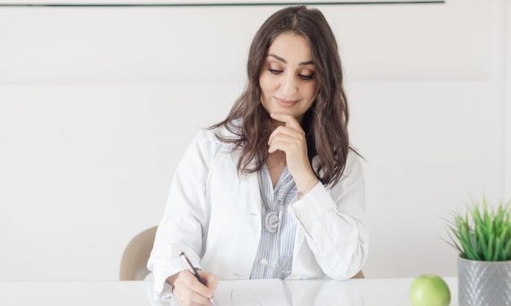 A female nutritionist in a lab coat writing notes with an apple and plant on the desk.
